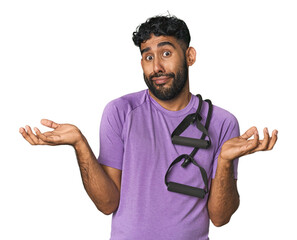 Hispanic male in sportswear with bands in studio doubting and shrugging shoulders in questioning...