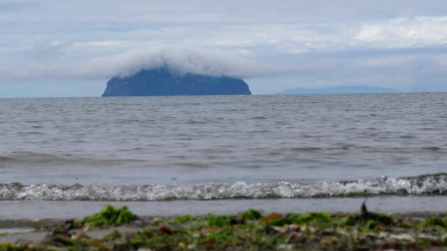 Mysterious round shape island with cloud cap on top, ground level shot of edge of beach, calm waves and round shaped Ailsa Craig island in Firth of Clyde.