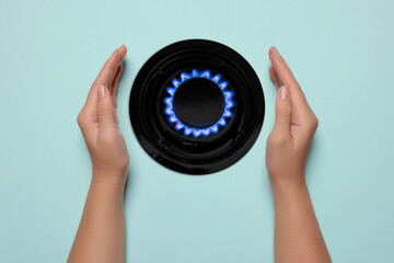 Woman protecting gas burner with blue flame on light blue background, top view