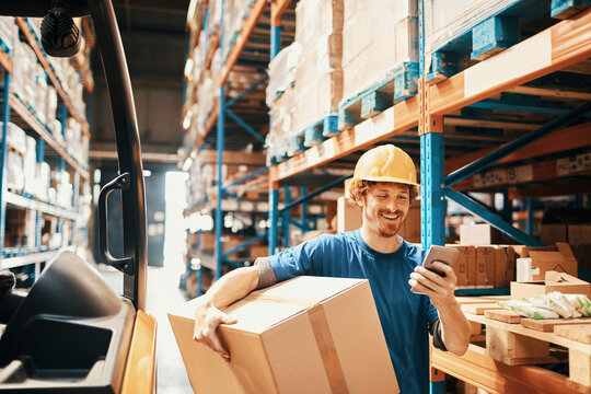 Smiling male worker using smartphone and carrying box in large warehouse