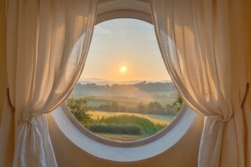 Elegant linen curtains on a circular window, capturing a tranquil view of a sunrise over the countryside.