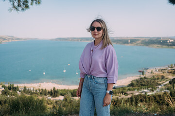 A girl in a lilac blouse and sunglasses on the background of an azure pond. Zhinvali reservoir, pine and tourist