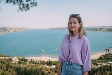 A girl in a lilac blouse and sunglasses on the background of an azure pond. Zhinvali reservoir, pine and tourist