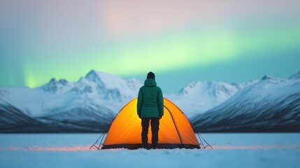 A solitary figure stands before a bright orange tent, surrounded by snow-covered mountains and illuminated by the vibrant colors of the northern lights.