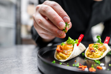 A close-up of a hand garnishing shrimp tacos with fresh herbs. The tacos are served on a black plate, showcasing vibrant ingredients, perfect for food-related content.