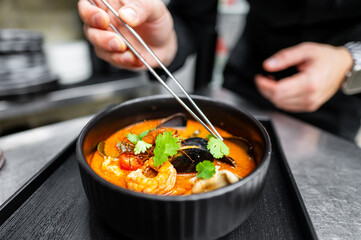 A chef expertly uses chopsticks to garnish a bowl of vibrant seafood stew, featuring shrimp, mussels, and fresh herbs. The setting showcases a professional kitchen ambiance.