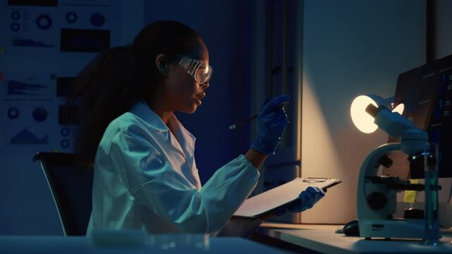 Young female scientist wearing lab coat, gloves and protective glasses taking notes on clipboard and observing samples under microscope in modern laboratory at night