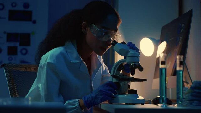 Young female scientist wearing lab coat and goggles using microscope in modern laboratory at night, illuminated by desk lamp and computer screen