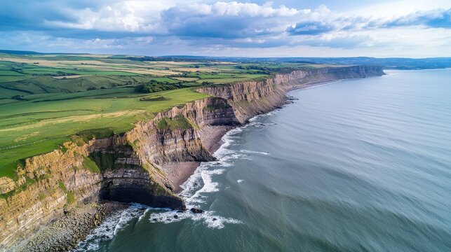 Aerial view of dramatic coastal cliffs meeting the tranquil ocean under a summer sky adorned with clouds and lush green hills nearby