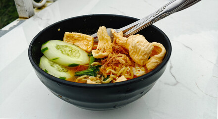 The bowl contains cooked noodles in a broth, garnished with slices of cucumber, fried shallots, and crispy crackers.