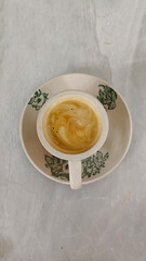 The image shows a cup of coffee with a creamy froth on top, placed on a saucer. The cup and saucer are white with green floral patterns.