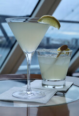 two pale yellow cocktails in front of a cruise ship window with a bokeh view of the shore