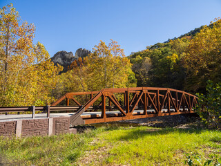 A rusty iron bridge with Seneca Rocks in the background behind trees in fall colors and a blue sky...