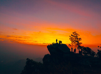Obraz premium Silhouette of a group of people climbing on a large cliff. Colorful sky of orange, yellow, pink, and blue in the morning at sunrise. There are mountains and trees around with copy space on top.