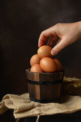 Hand holding brown egg over the round wooden bowl full of brown eggs, perfect for Easter on dark background 