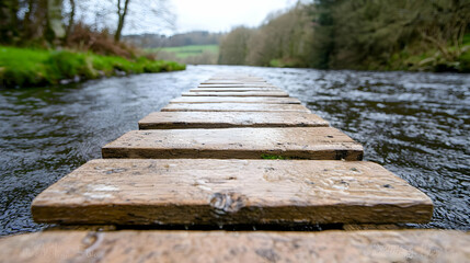 Fototapeta premium Wooden footbridge extends over a flowing river in a lush green forest during a cloudy day.