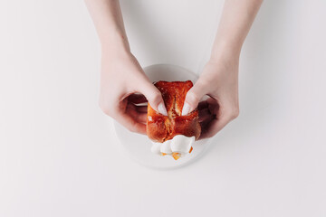 Woman's hands tearing, unwrapping, and crushing a pastry on a white plate against a white background