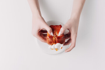 Woman's hands tearing, unwrapping, and crushing a pastry on a white plate against a white background