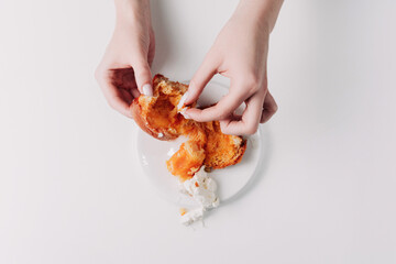 Woman's hands tearing, unwrapping, and crushing a pastry on a white plate against a white background