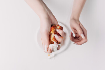 Woman's hands tearing, unwrapping, and crushing a pastry on a white plate against a white background