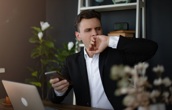 A young man in a black suit yawns while holding a smartphone in a modern office. The workspace includes plants, a laptop, and an overall relaxed, professional atmosphere.