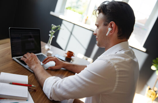A man is sitting at a modern desk, working on a laptop. He is wearing white wireless earbuds and writing in notebook.Сup of coffee and a smartphone are placed on the desk in well-lit office setting.