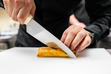 A chef's hands skillfully slice a golden, crispy spring roll on a white cutting board, showcasing culinary expertise in a professional kitchen setting.