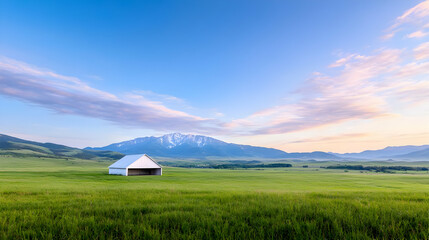 Obraz premium White barn stands in a vast green meadow under a colorful sunrise sky with mountains in the background.