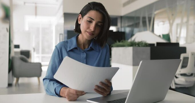 Latina woman, office employee sit at desk reviewing document using laptop, analyzing data, drafting report, cross-checking information in contract, working on financial invoices, receipt, or statement