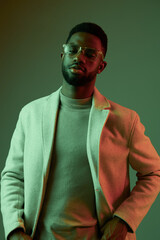Stylish young man wearing glasses and a light coat against a colorful background, showcasing confidence and modern fashion trends with green and red lighting