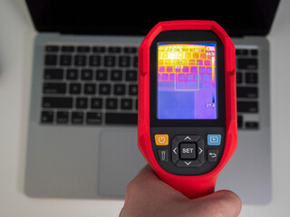 Technician holding thermal imaging camera, scanning laptop surface, detecting heat signatures and potential thermal anomalies during professional diagnostic maintenance process