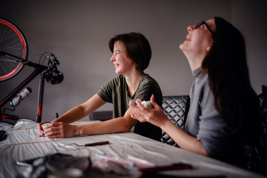Two women sharing a laugh while working on a bicycle indoors. The scene is lively and cheerful, highlighting teamwork and enjoyment in a casual setting.