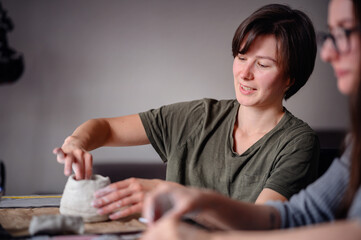Two women engaged in crafting clay pottery together. One woman shapes a clay piece while the other...