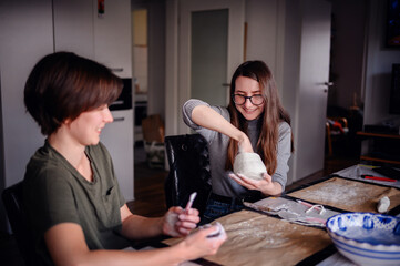 Two women engaged in a clay sculpting project, conversing and sharing a creative moment. The image captures collaboration and artistic expression in a relaxed setting.