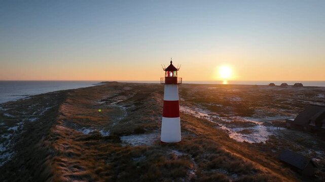 Aerial orbit of red and white striped lighthouse situated on a coastal landscape at sunrise in winter. Lighthouse West-List on the Ellenbogen (the "Elbow"), the peninsula on northern coast of Sylt.