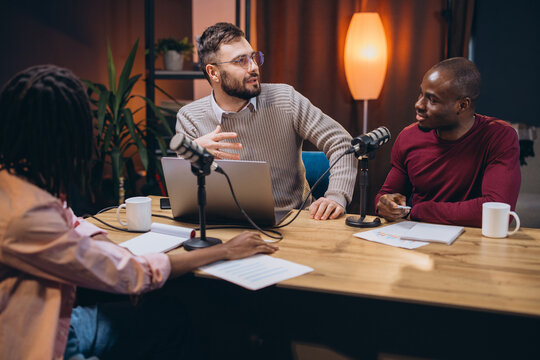 Three podcasters discussing a new topic in a professional home studio, using microphones and laptop