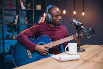 Young African American musician performing acoustic guitar, singing into microphone during home...