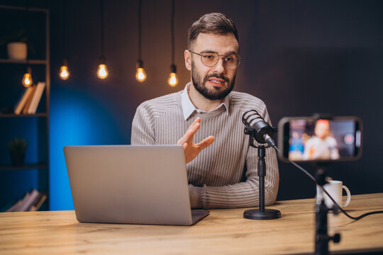 Young man recording a podcast using a professional microphone and laptop, talking and gesturing while looking at the camera