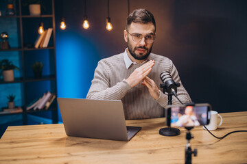 Young man gesturing while recording a podcast and live streaming using professional microphone and smartphone in home studio