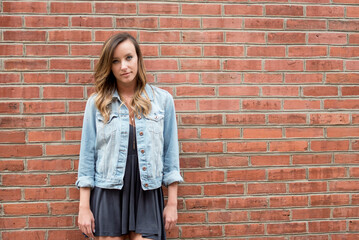 Beautiful young woman poses wearing short black dress and denim jacket standing in front of brick wall