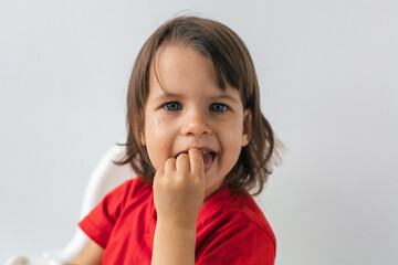 Curious little girl with dark hair wearing a red shirt, eating with her fingers and looking at the camera with food crumbs on her face. Concept of childhood, exploration, and natural eating