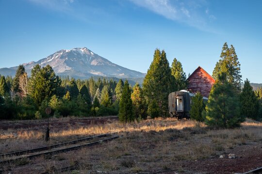 Abandoned train car on tracks, nestled amongst trees and a red barn, with a snow-capped Mount Shasta in the background. Tranquil rural scene. , Mccloud, California, USA