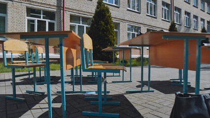Empty desks and chairs stand outside a school building on a sunny school day, suggesting an outdoor class or break time