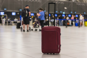 Red-brown rolling travel suitcase at the airport terminal with blurred passengers in the background. Journey, luggage, and aerodrome concepts.