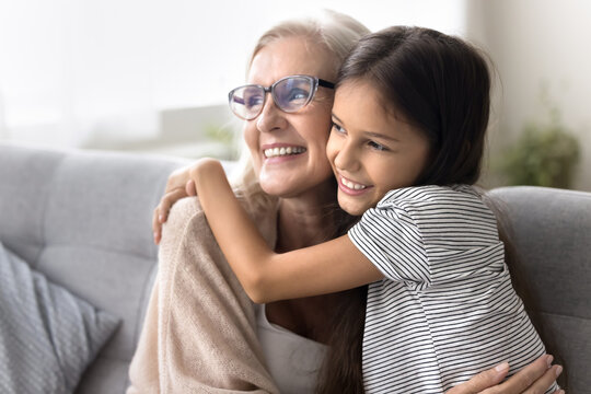 Dreamy aged single mom little preteen age daughter cuddle on couch at home look away imagine better future visualize plans. Happy elder nanny hug beloved child ward enjoy tender sweet moments together