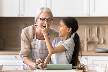 Playful small tween age girl kidding joking in house kitchen with laughing senior grandmother put...