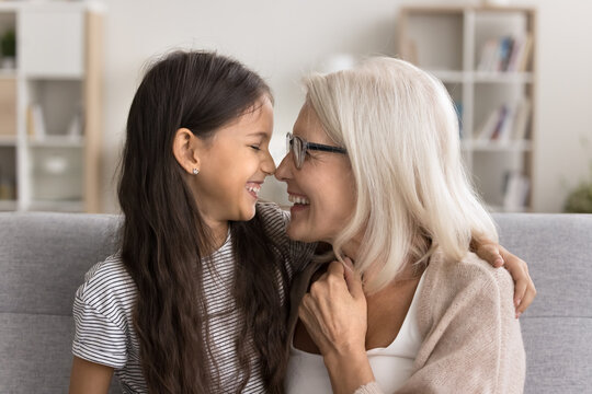Adorable moment. Cute little 10 years old girl tender touching noses with smiling affectionate older age grandmother. Happy loving granny granddaughter playing on cozy sofa cuddling feel warmth unity