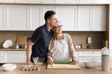 Dreamy young son tender hug shoulders of smiling retired mum baking buns at domestic kitchen look...