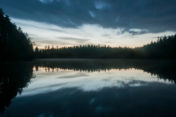 A scenic and calm lake view at night with mist and reflected forest on the surface in Finland