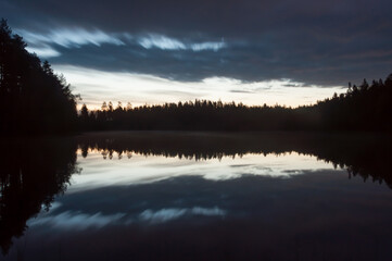 A scenic and calm lake view at night with mist and reflected forest on the surface in Finland